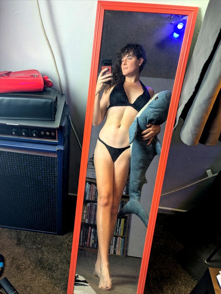 A woman in a bikini takes a mirror selfie with her stuffed ikea shark toy. She has very nice abs.