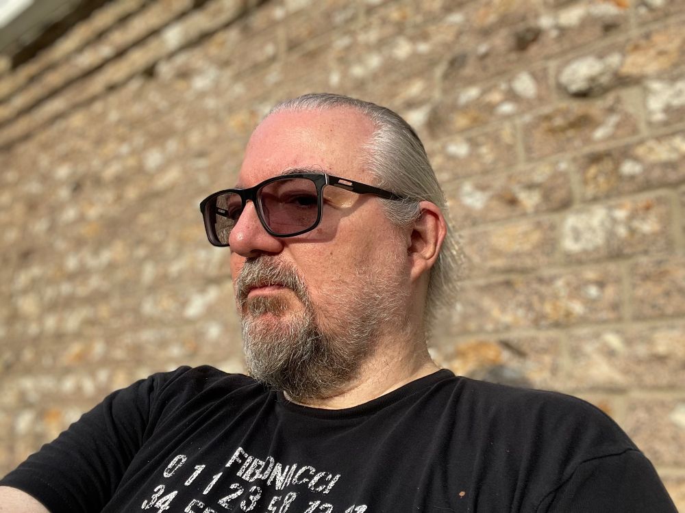 me looking entirely too serious sitting outside in the sun with dark glasses and a black shirt with a beige brick wall behind me.