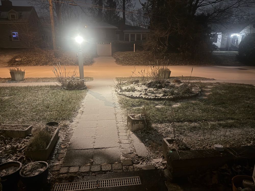 Night-time snow storm pic: A powdering of snow on a front yard walk with a poke-light and street, with glowing houses across the street