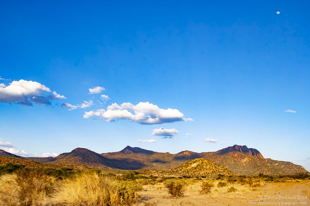 Arid hills bathed in an orange glow from the setting sun and a deep blue sky above them with just a few fluffy white clouds