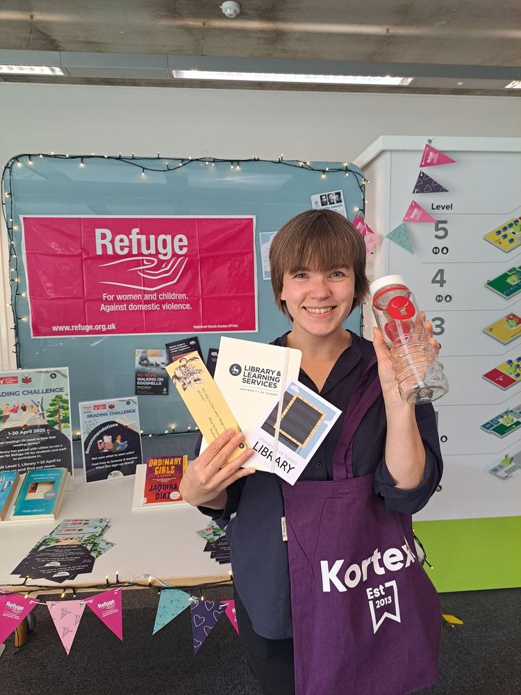 Librarian holding her prizes from the Reading Challenge. She is holding a Kortext purple bag, water bottle with red stag head logo, notebook, bookmark, and standing in front of the Refuge charity display in the Library.