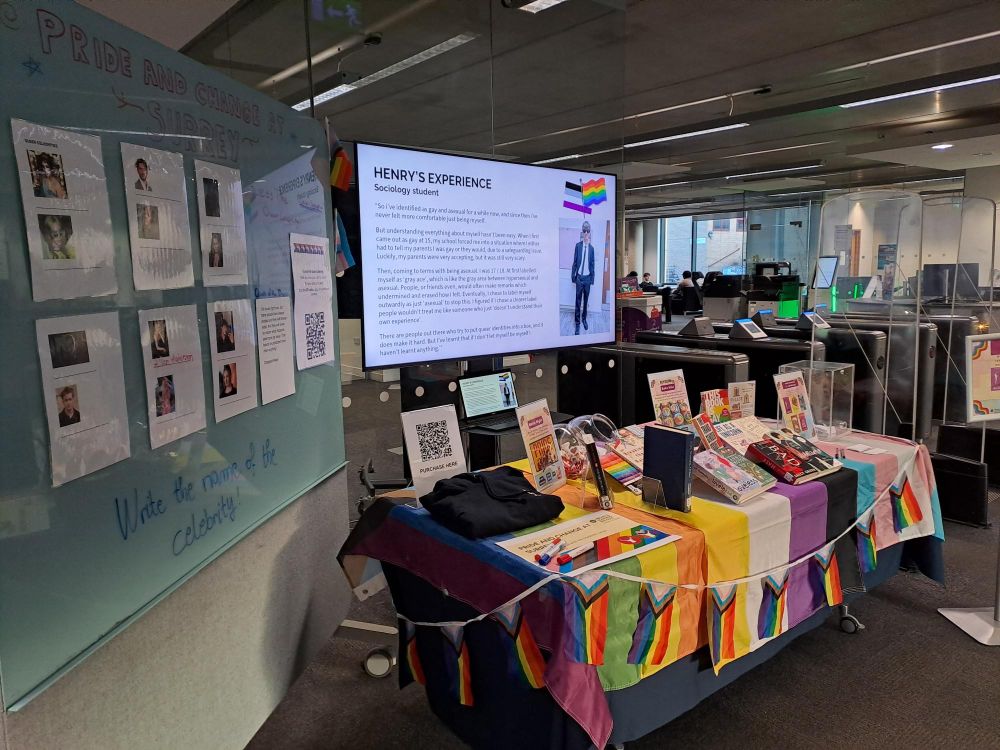 Display of LGBTQ+ books on the table atop LGBTQ+ and Pride flags and rainbow bunting. The screen is showing student named Henry's photo and his blog. On the side, to the left, is a big white board where student can guess the names of the queer celebrities on the board. 