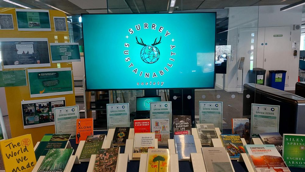 Image shows the Student curator corner display. In the background is logo of the Sustainability society on the screen - it is a globe with two black antlers that represent the stag, which is the University mascot, poking out at the top. In the forefront is a display of the collection of recommended books and the yellow writing board with photos of the society and movie screening times as well as information about the green pledge. 