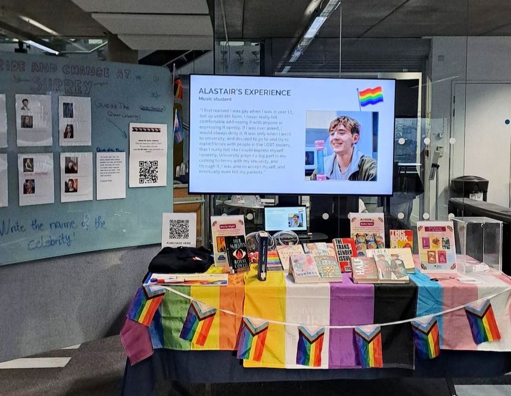 Display of LGBTQ+ books on the table atop LGBTQ+ and Pride flags and rainbow bunting. The screen is showing the student, Alastair's photo and his blog. On the side, to the left, is a big white board where student can guess the names of the queer celebrities on the board. 