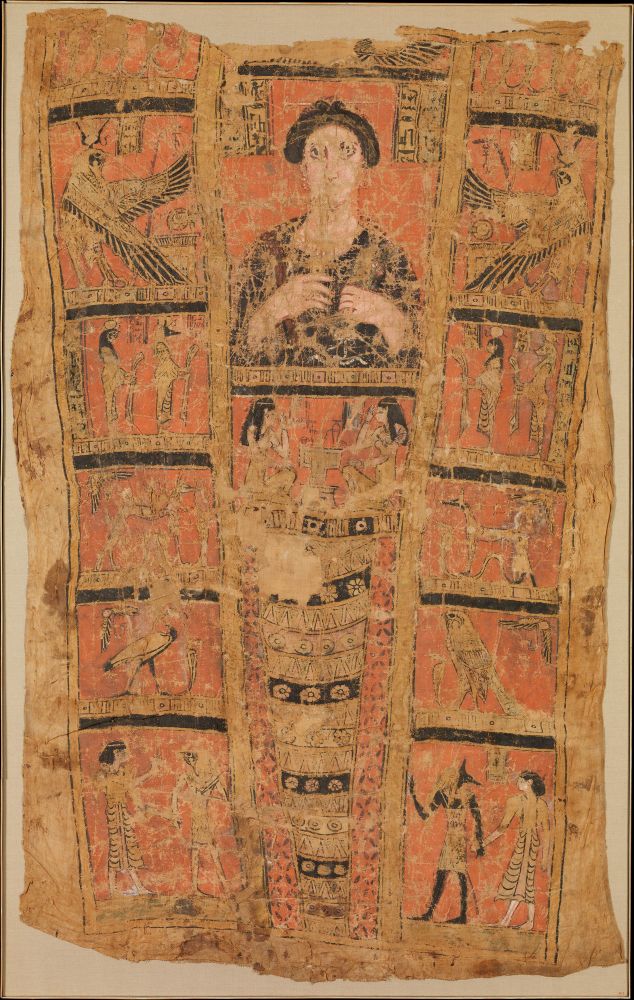 Description adapted from The Met: “Placed over the supine mummy the naturalistic painted bust would lie over the face, and the panels of gods would have been draped along its sides. The woman’s rosy face and large eyes are striking. A long bone or ivory pin holds coils of her hair in place on top of her head, and she holds a small wreath in one hand. Over her abdomen Isis and Nephthys mourn, and over her lower body are placed the long strands of the late protective ornament as well as a net garment. Columns on either side of her head are invocations to the god Osiris, but her name is lost. The side panels are paired, showing the goddesses Isis and Nephthys as red kites (a bird related to the hawk); the four mummy-shaped “sons of Horus” associated with the bodily integrity; jackals associated with transit and cemetery; the Thoth-ibis and the Horus-falcon; and at the bottom scenes of the dead woman led by Horus and alternatively Anubis into the afterlife.“