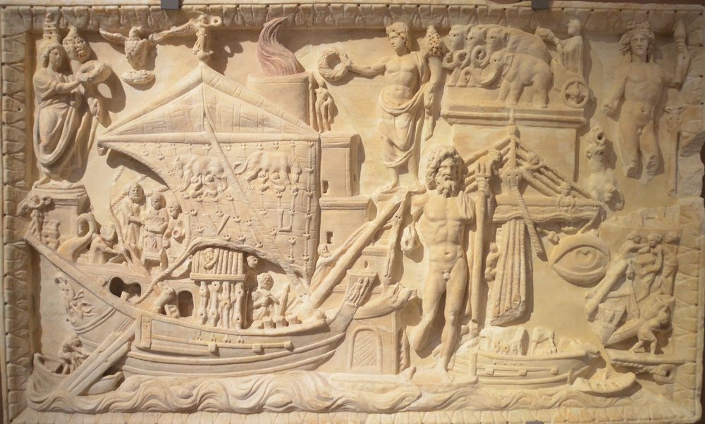 Image description, modified from Ostia-Antica.org: the relief depicts waves which carry two ships. In the top left corner is a female figure. She holds a cornucopiae in her left hand and a wreath in her right hand. On her head is a lighthouse similar to other depictions of the lighthouse of Portus, suggesting that the figure is the personification (Genius) of Portus. To the right of the figure flies an eagle with outstretched wings. With his claws he holds a wreath. Further to the right, is the lighthouse of Portus. On the third upper floor of the lighthouse is a male, naked (bearded?) figure. Further to the right is a male figure standing on a square pedestal, facing left. With his left hand he holds a cornucopiae. With his right hand he holds a wreath over the head of the figure standing on the lighthouse. A cargo ship occupies most of the left half of the relief. Romulus and Remus can be seen suckled by the she-wolf, facing left and right respectively. On top of the main mast is a winged Victoria.