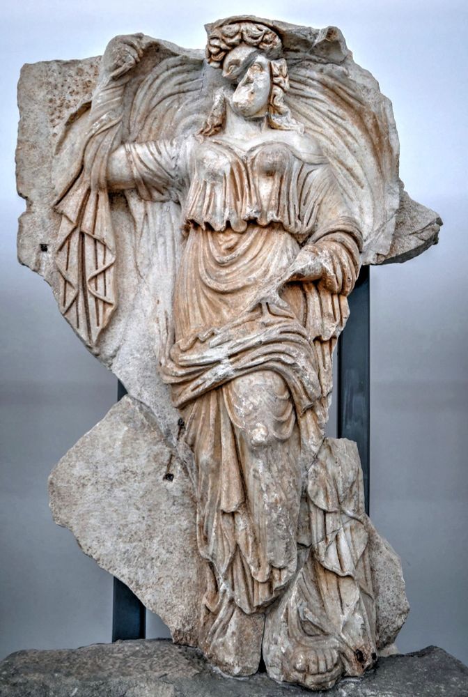Image description: A detailed relief of Hemera standing front on to the viewer. What she held in her left hand is unclear. In her right hand she holds up the billowing folds of her gown. Her hair sits down to her shoulders and there’s damage to her face obscuring her original features. Held by the Aphrodisias Museum.