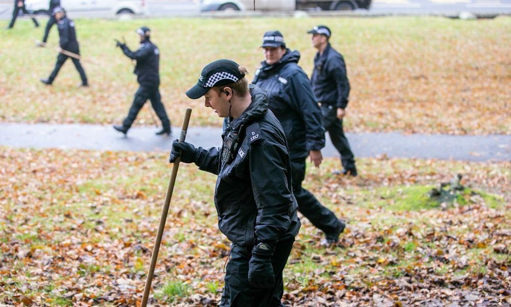 A row of police officers walk in a line across a field, searching for evidence