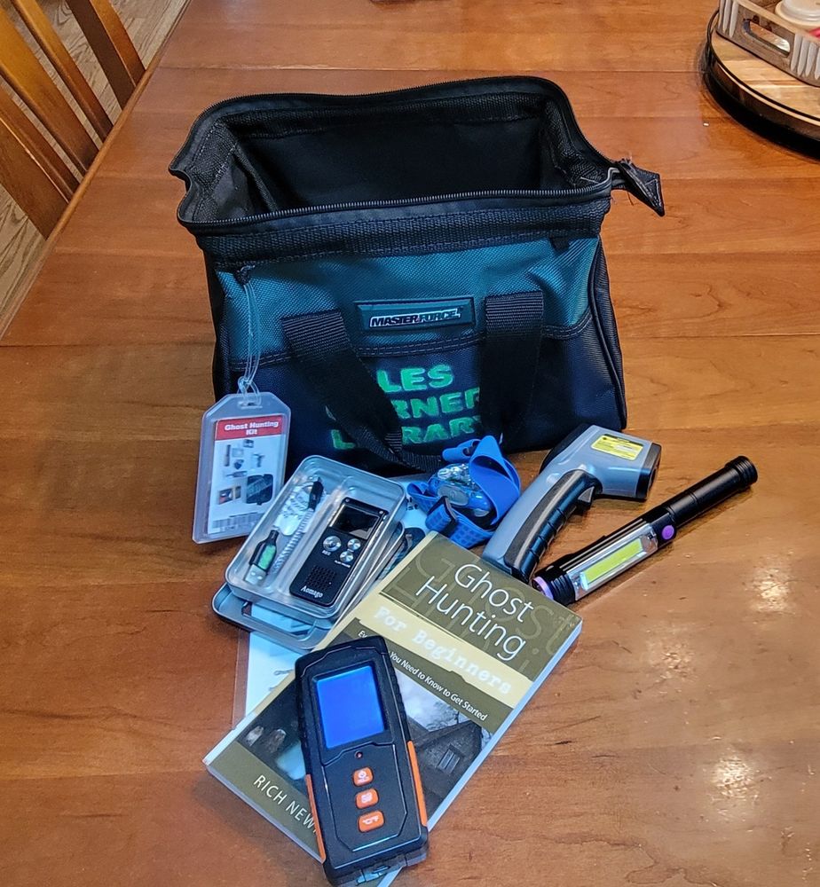 Wood table with a small satchel unpacked to show the contents of a ghost hunting kit from the Hales Corner public library, comprising an EMF reader, UV flashlight, digital voice recorder, head lamp, digital temp gauge with laser targeting and a copy of 