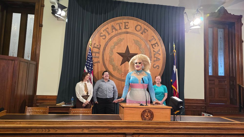 Maxine LaQueen is a trans woman drag queen in a sparkly trans pride flag dress and a huge blonde hairdo. She's speaking in front of the seal of the state of Texas at a press conference, with other trans and queer Texans behind her on the platform. 
