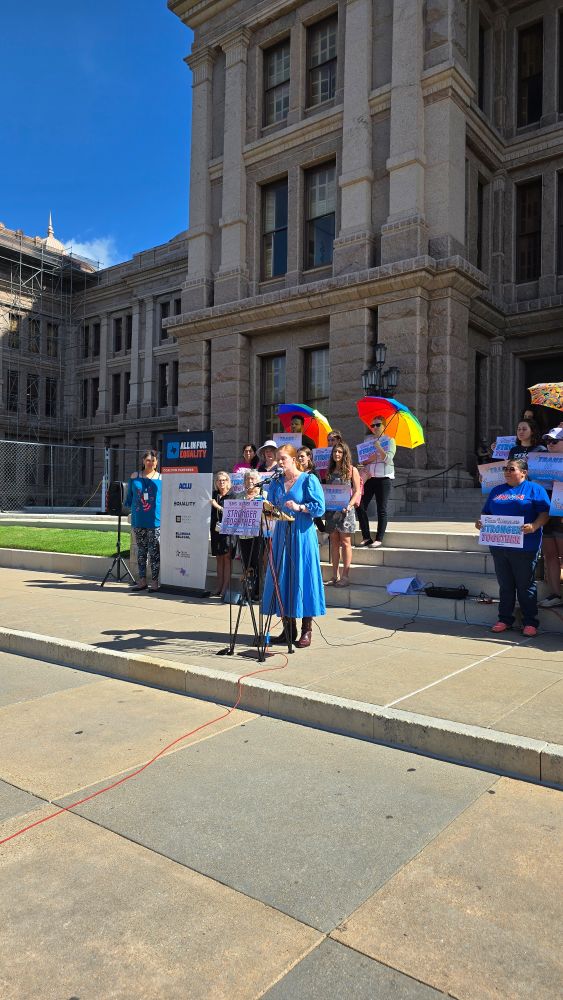 Sen. Molly Cook speaking at the microphone at the Texas Capitol steps, wearing a long blue dress. 