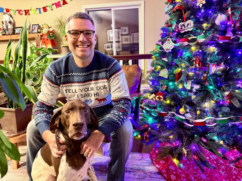 FiniteSingularity and Ollie the German Shorthair Pointer sitting next to a Christmas tree.  Finite is wearing a “Tell Your Dog I Said Hi” Christmas Sweater