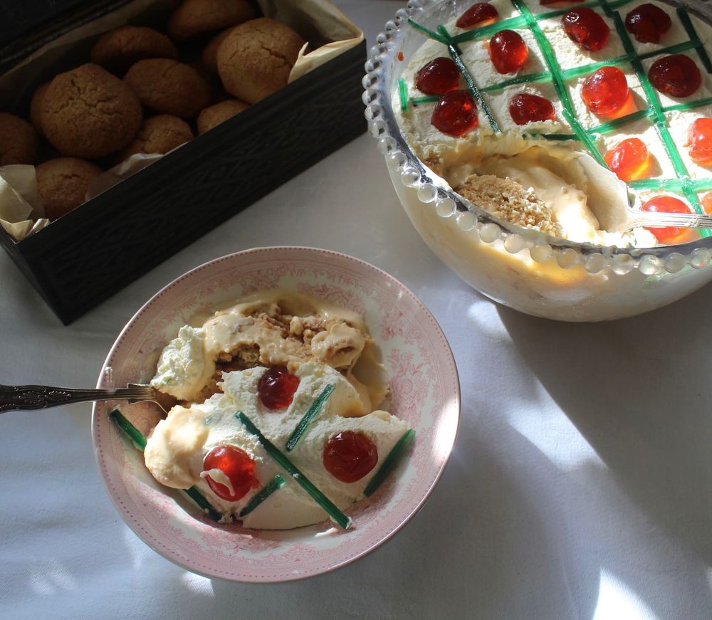 Glass bowl of trifle decorated with glace cherries and Angelica. Pink dessert bowl of trifle with silver spoon.  Box of ratafia biscuits.  Damask white tablecloth.