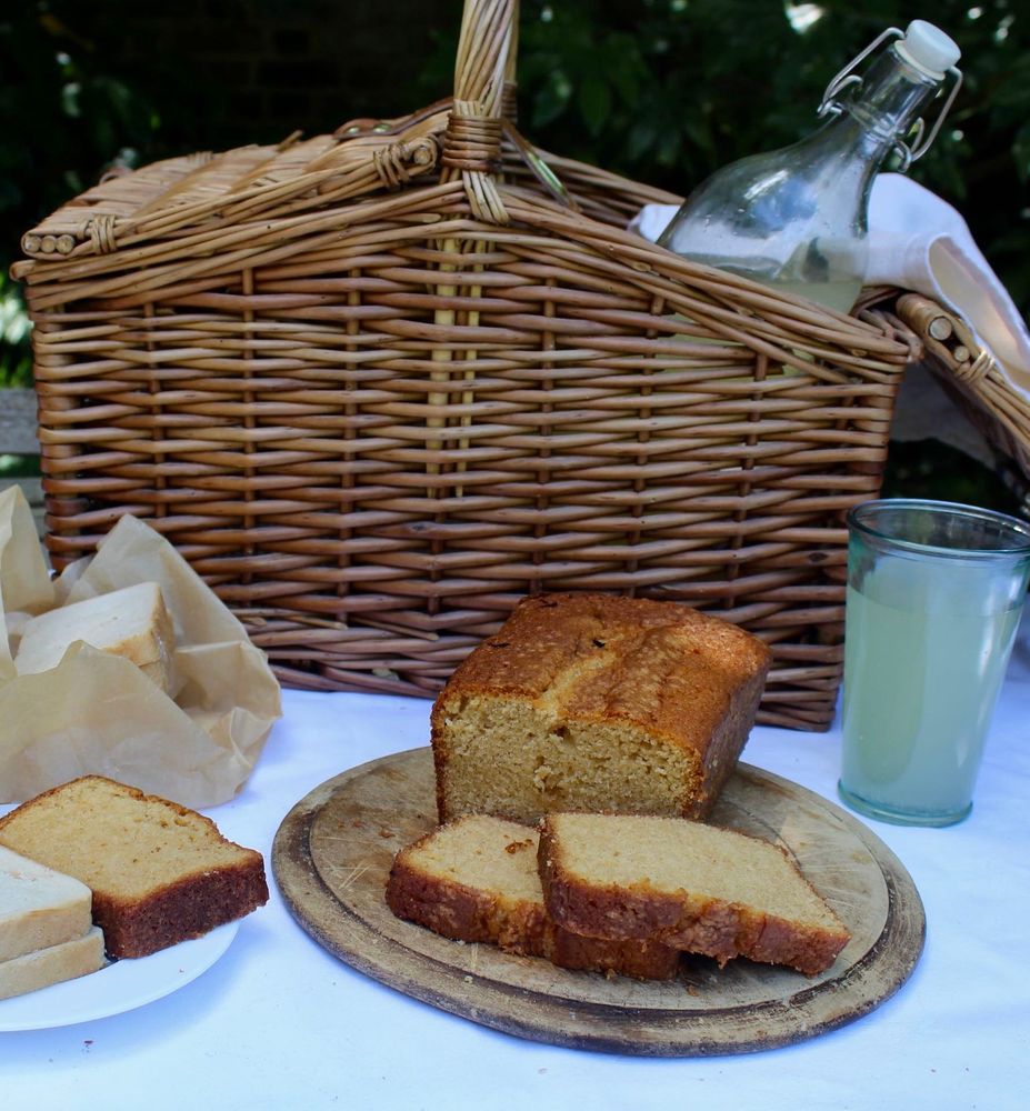 Pound cake on wooden board on white tablecloth surrounded by plate with sandwich and slice of cake, sandwiches in greaseproof paper, picnic hamper with bottle of ginger beer and glass of ginger beer.
