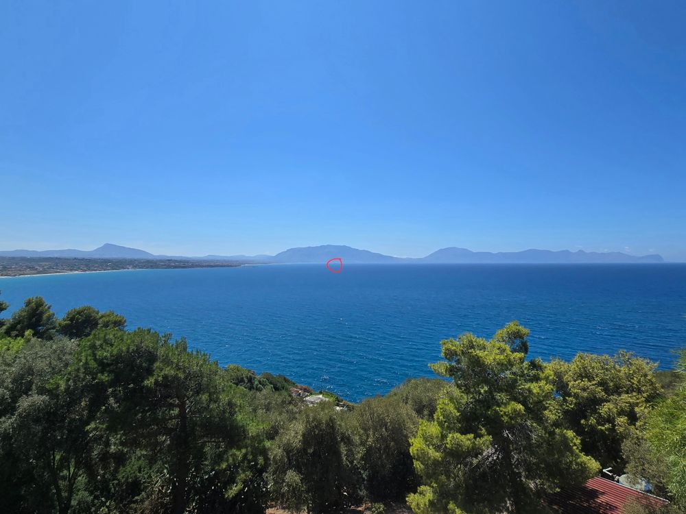 Panoramaaufnahme einer mediterranen Küstenlandschaft mit tiefblauem Meer, bewaldeten Hängen im Vordergrund und einer weitläufigen Gebirgskette am Horizont. In der Bildmitte ist ein kleiner roter Kreis markiert, vermutlich zur Hervorhebung eines kaum sichtbaren Objekts auf dem Wasser. Der Himmel ist klar und wolkenlos.