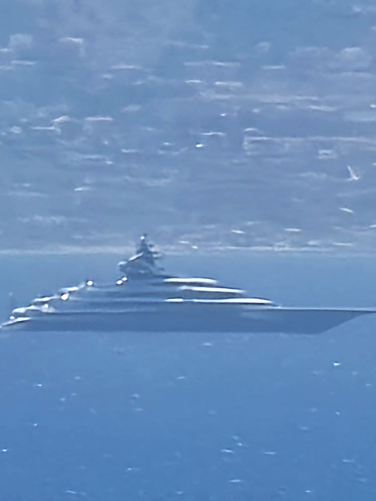 Unscharfe Teleaufnahme einer luxuriösen Superyacht, die ruhig auf dem tiefblauen Meer vor einer felsigen, sonnenbeschienenen Küste liegt. Die Yacht wirkt durch die Perspektive und Lichtreflexion fast schwebend. Im Hintergrund sind verschwommene Terrassenhänge oder Gebäude erkennbar.