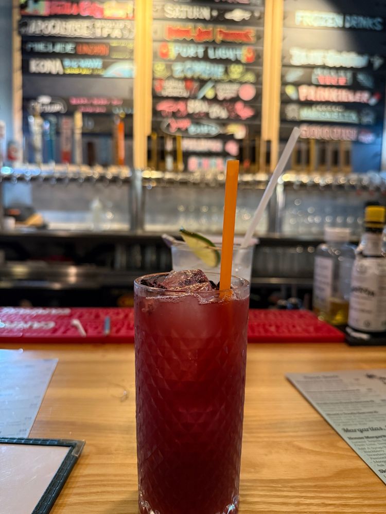 A dark cocktail in a tall skinny glass, with an out-of-focus background of a chalkboard drink menu. 