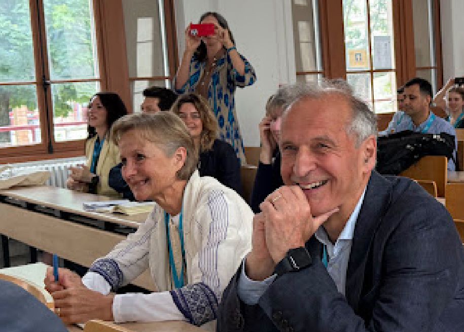 Roger Matthews (looking surprised) sits next to Wendy Matthews at a desk in front of a room of people who were clearly in on the conspiracy 