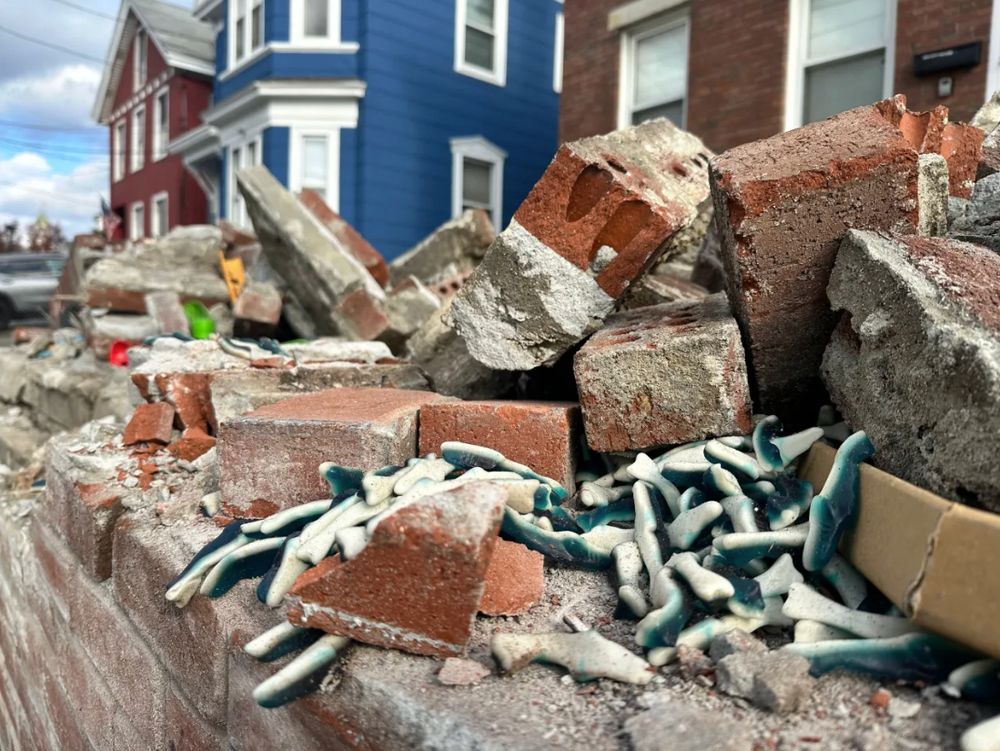 A photograph of bricks and rubble with a large pile of blue and white gummy sharks sprinkled into it. Photo credit to Aaron Curtis/Lowell Sun. Photo appeared with this story: https://www.lowellsun.com/2025/11/12/not-so-sweet-tractor-trailer-hauling-candy-cargo-flips-at-end-of-lowell-connector/