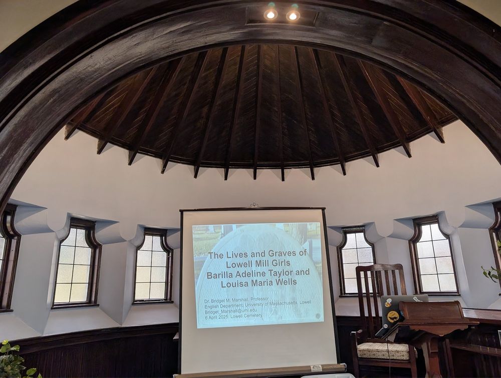 Photo of a room with a screen in front projecting slides for "The Lives and Graves of Lowell Mill Girls Barilla Adeline Taylor and Louisa Maria Wells."