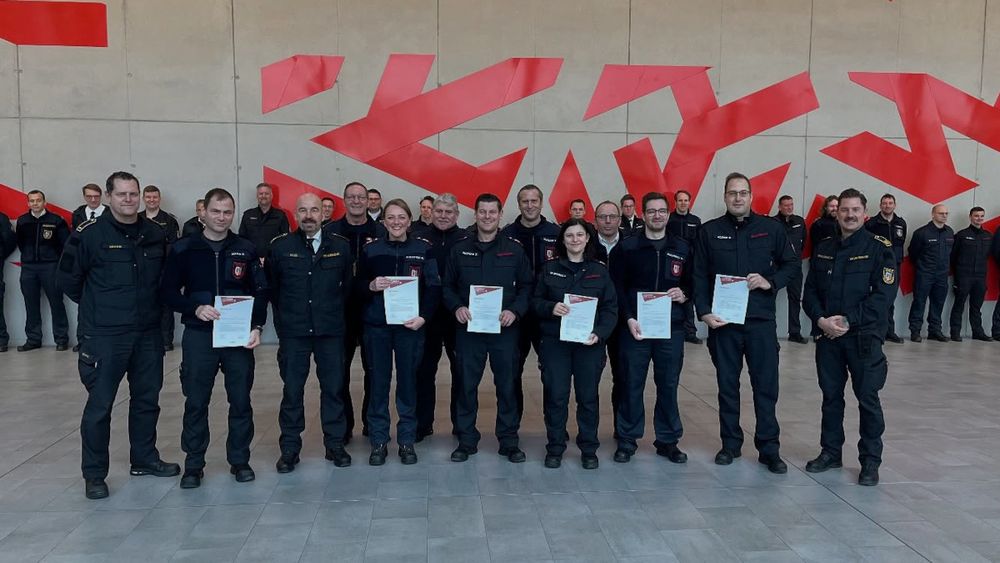 Gruppenfoto von Einsatzkräften der Feuerwehr in Uniform vor einer Wand mit großen roten Pfeilformen. In der vorderen Reihe stehen mehrere Feuerwehrangehörige und halten Urkunden in den Händen, dahinter sind weitere Kolleginnen und Kollegen aufgestellt. Das Bild zeigt die feierliche Übergabe von Abschlussurkunden in einer modern gestalteten Halle.