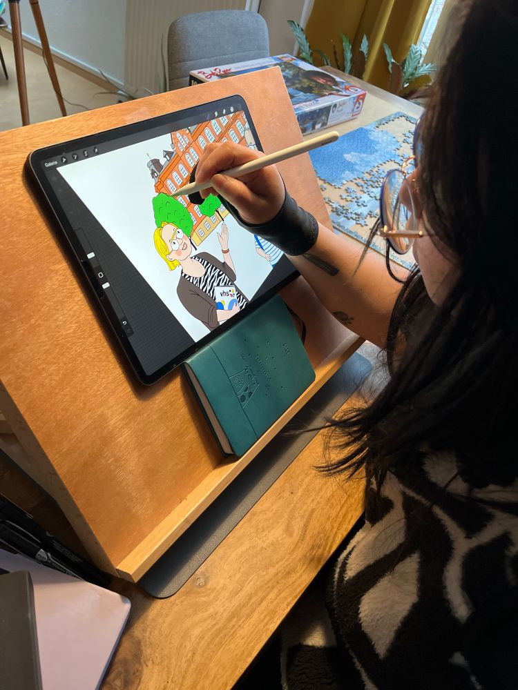 Die Illustratorin Nontira Kigle sitzt an einer Tischstaffelei, auf dem ein iPad ist und zeichnet darauf eine fröhlich bunte Illustration für Kinder. Auf dem Tisch ist ein angefangenes Puzzle von einem Bob Ross Gemälde.
