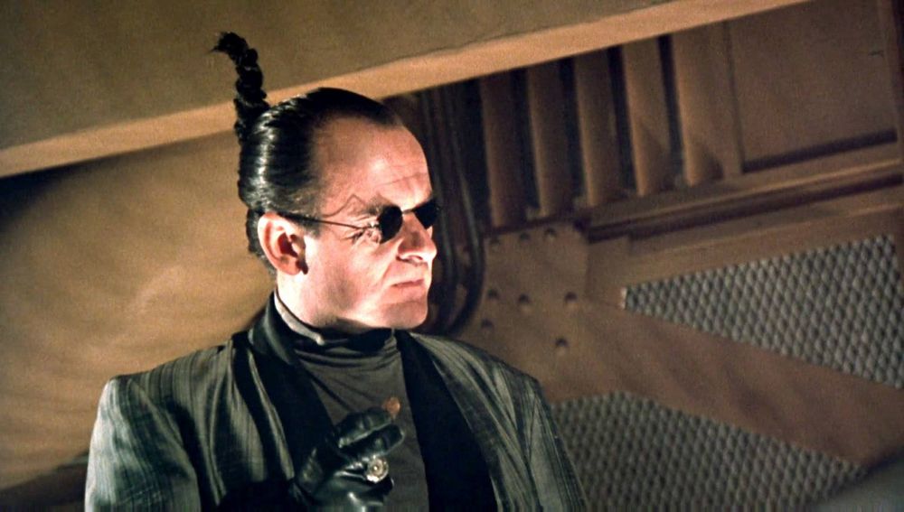 Joe Pesci in MOONWALKER, with a ponytail sticking straight up, wearing small round sunglasses, a black jacket and black turtleneck