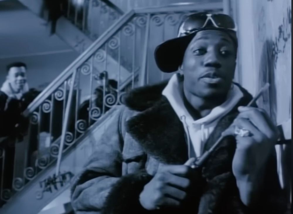 A black and white shot from the short film "Bad," with Wesley Snipes in the foreground in front of an apartment stairwell, wearing a leather bomber jacket with fur over a white hoodie and a baseball cap worn sideways. He's holding a scraping tool.