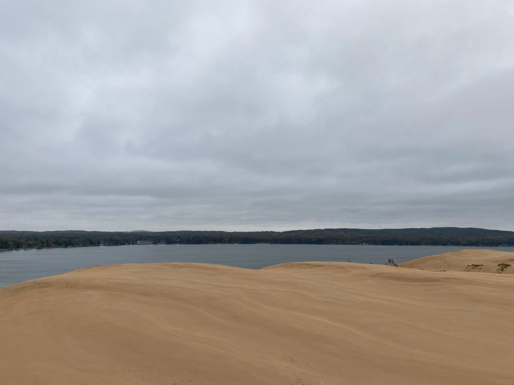 Wind blown sand dunes in the foreground looming high over a forest wrapped lake under grey autumn skies
