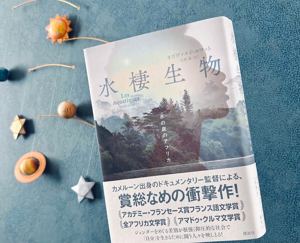 惑星のモビールが掛かった青い壁の前で『水棲生物　水の底のアフリカ』オズヴァルド・ルワット／大林薫訳（講談社）を持っている写真。
本の帯には「カメルーン出身のドキュメンタリー監督による、賞総なめの衝撃作！ジェンダーをめぐる差別が根強く抑圧的な社会で「自分」を生きるために闘う人々を映しとる！」と書かれている。