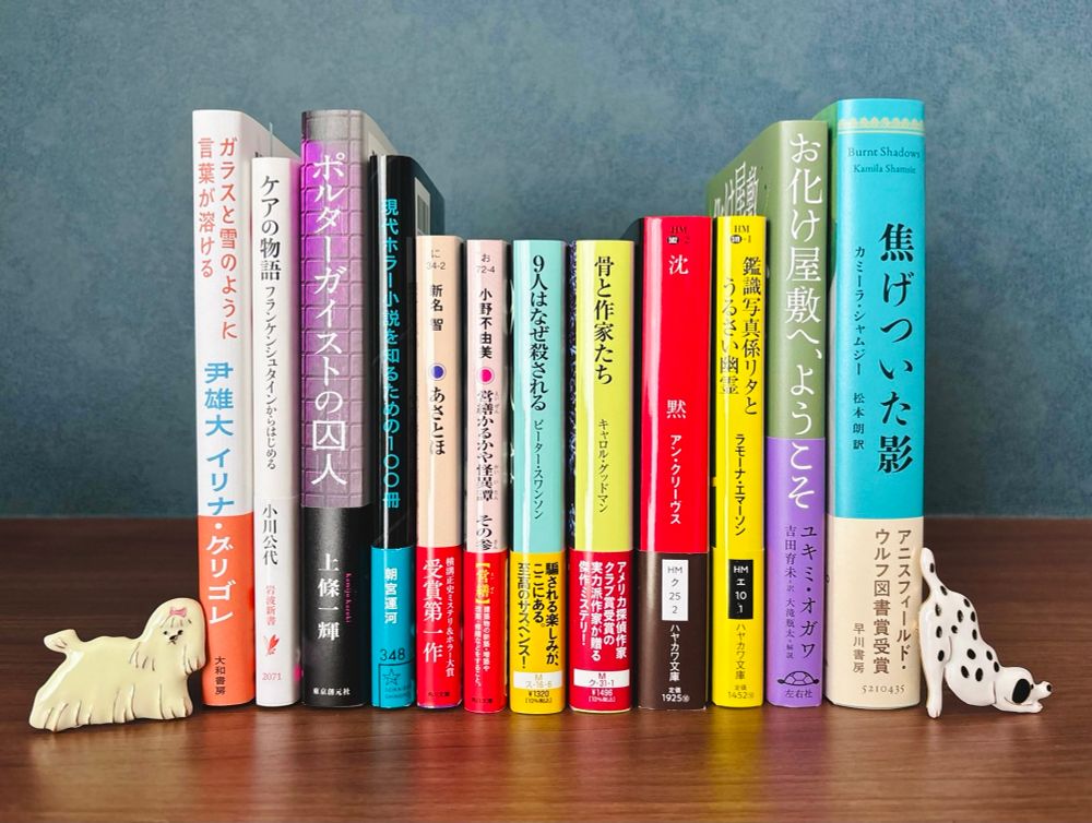 12冊の本が棚の上に、背表紙を向けて立てて並べて置かれた写真。右側から順に、
◆『焦げついた影』カミーラ・シャムジー／松本朗訳（早川書房）
◆『お化け屋敷へ、ようこそ』ユキミ・オガワ／吉田育未訳（左右社）
◆『鑑識写真係リタとうるさい幽霊』ラモーナ・エマーソン／中谷友紀子訳（ハヤカワ文庫）
◆『沈黙』アン・クリーヴス／高山真由美訳（ハヤカワ文庫）
◆『骨と作家たち』キャロル・グッドマン／栗木さつき訳（創元推理文庫）
◆『9人はなぜ殺される』ピーター・スワンソン／務台夏子訳（創元推理文庫）
◆『営繕かるかや怪異譚　その参』小野不由美（角川文庫）
◆『あさとほ』新名智（角川文庫）
◆『現代ホラー小説を知るための100冊』朝宮運河（星海社）
◆『ポルターガイストの囚人』上條一輝（東京創元社）
◆『ケアの物語　フランケンシュタインからはじめる』小川公代（岩波新書）
◆『ガラスと雪のように言葉が溶ける　在日韓国人三世とルーマニア人の往復書簡』尹雄大／イリナ・グリゴレ（大和書房）