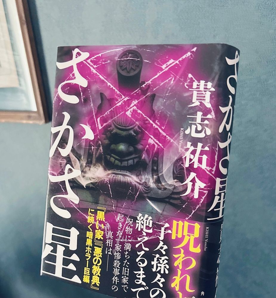 『さかさ星』を壁の前で持っている写真。本の帯には、「呪われよ　子々孫々の絶えるまで」「呪物に満ちた旧家で起きた一家惨殺事件の真相は──」と書かれている。