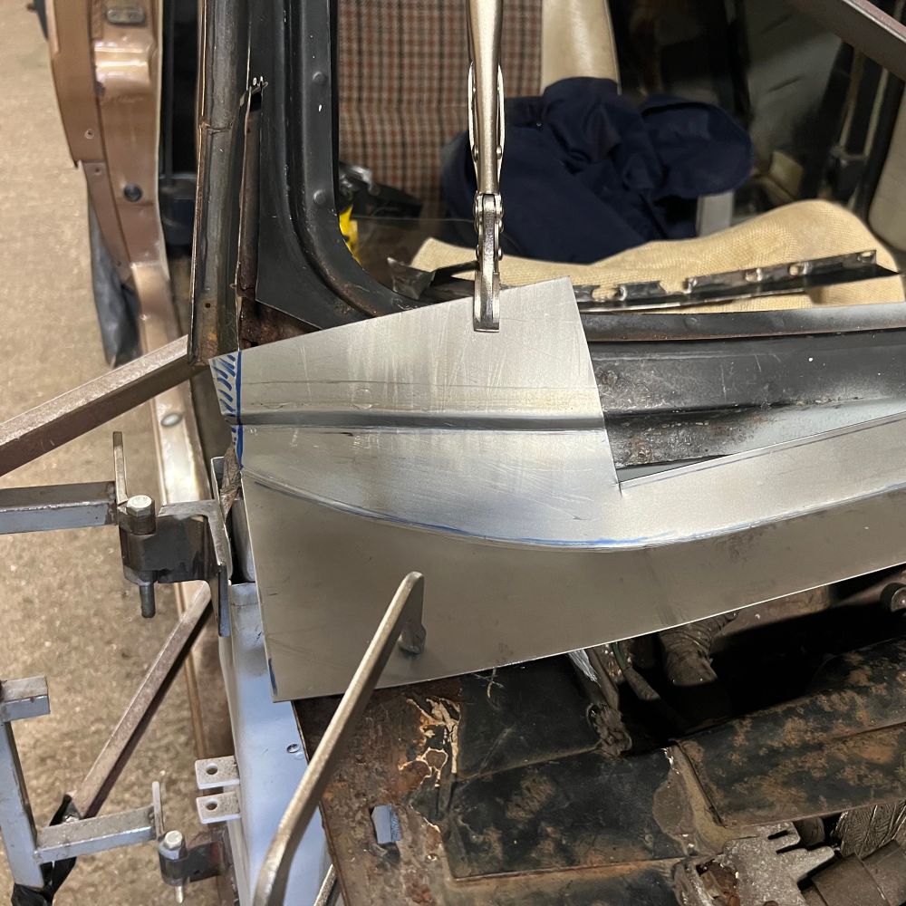 A piece of hand-shaped sheet steel clamped in place over the lower corner of the windscreen of a rusty car.