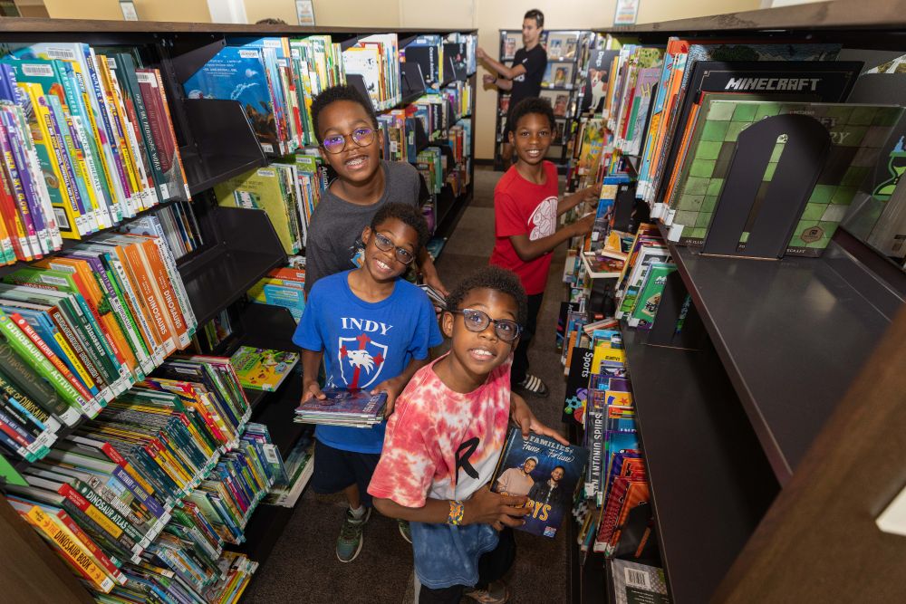 Group of  children smiling while looking for books in the bookshelves at the Fort Ben Branch Library. 