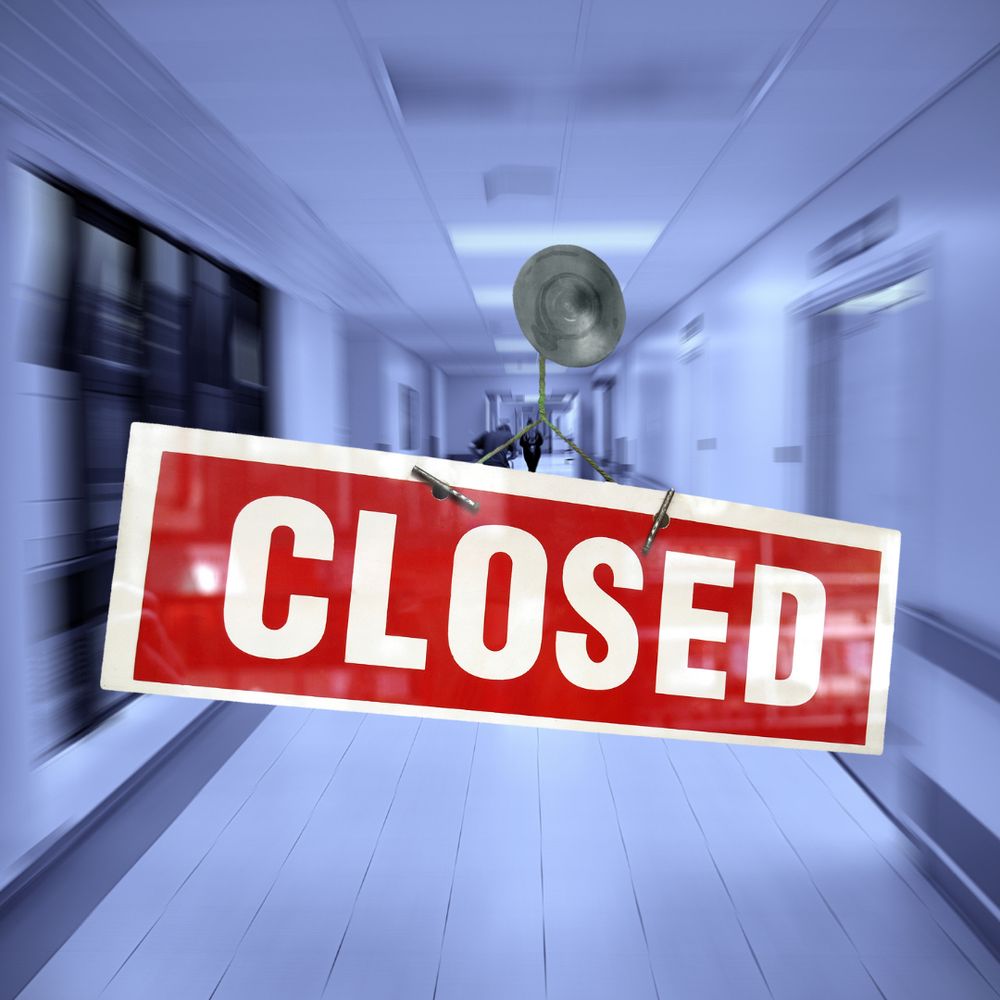 Grey image of a hospital corridor in the background over which is a red sign with white border and white writing saying CLOSED.