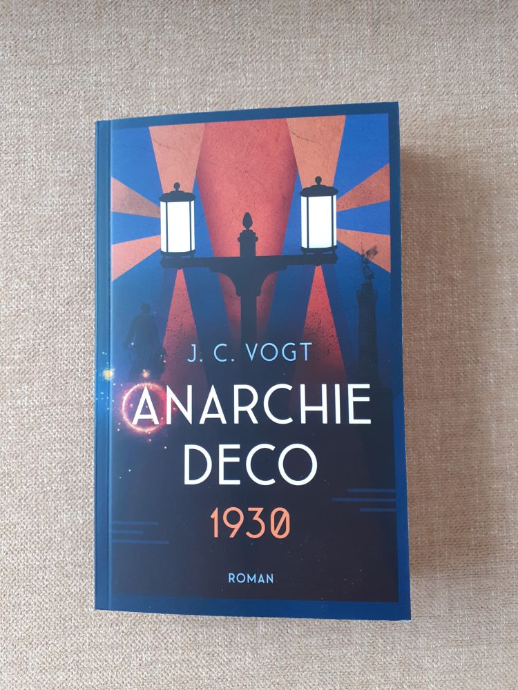 Foto: Auf beigefarbenem Hintergrund liegt der Roman "Anarchie Déco 1930" von Judith und Christian Vogt. Das Cover zeigt links die Silhouette einer Figur im Schatten zwischen den Lichtkegeln einer stilisierten zweiarmigen Straßenlaterne, rechts im Hintergrund die Berliner Siegessäule, und ist überwiegend in Orange- und Blautönen gehalten. Das Cover stammt von Sameena Jehanzeb (saje design).