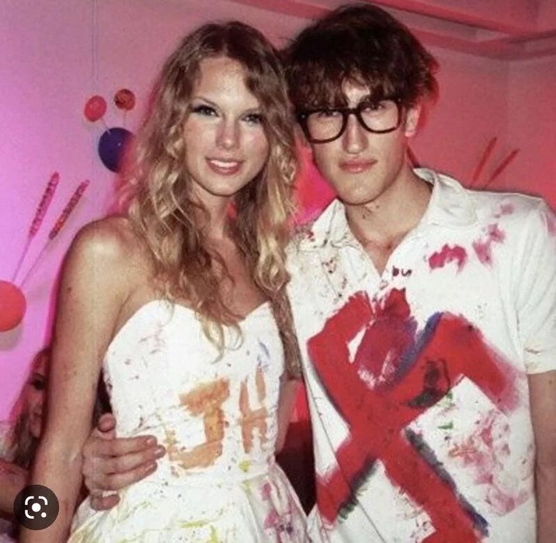 An older photo of Taylor Swift who was posing with a model at a Hollywood party. The guy has a giant red swastika on his shirt
