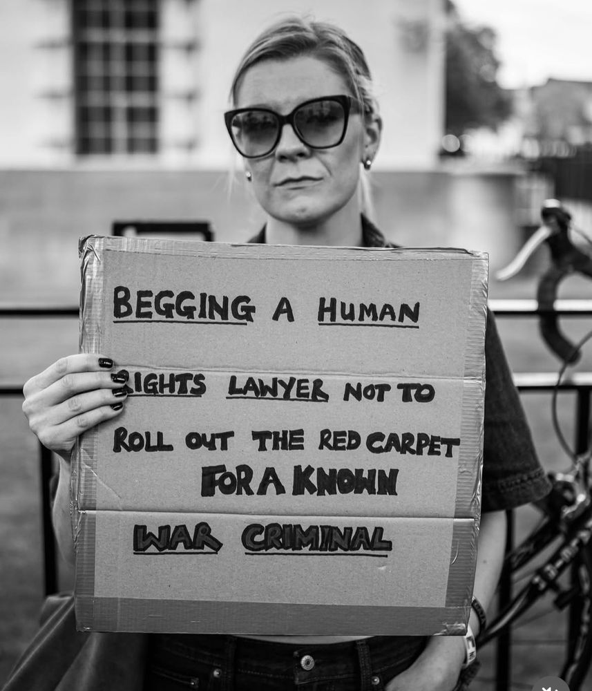 A woman holding up cardboard placard that reads ‘Begging a human rights lawyer not to roll out the red carpet for a known war criminal’