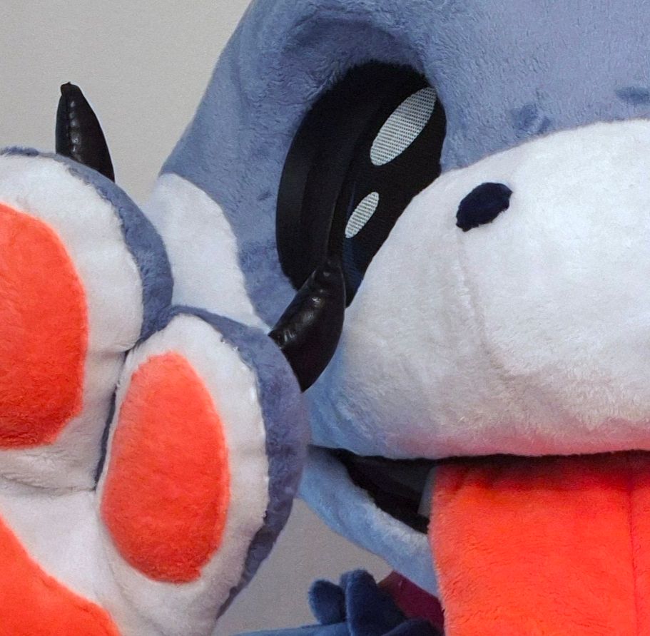 A cropped shot of a blue shark fursuit head, showing a big black eye, a round white snout  a big orange-pink tongue, and 2 digits of a paw