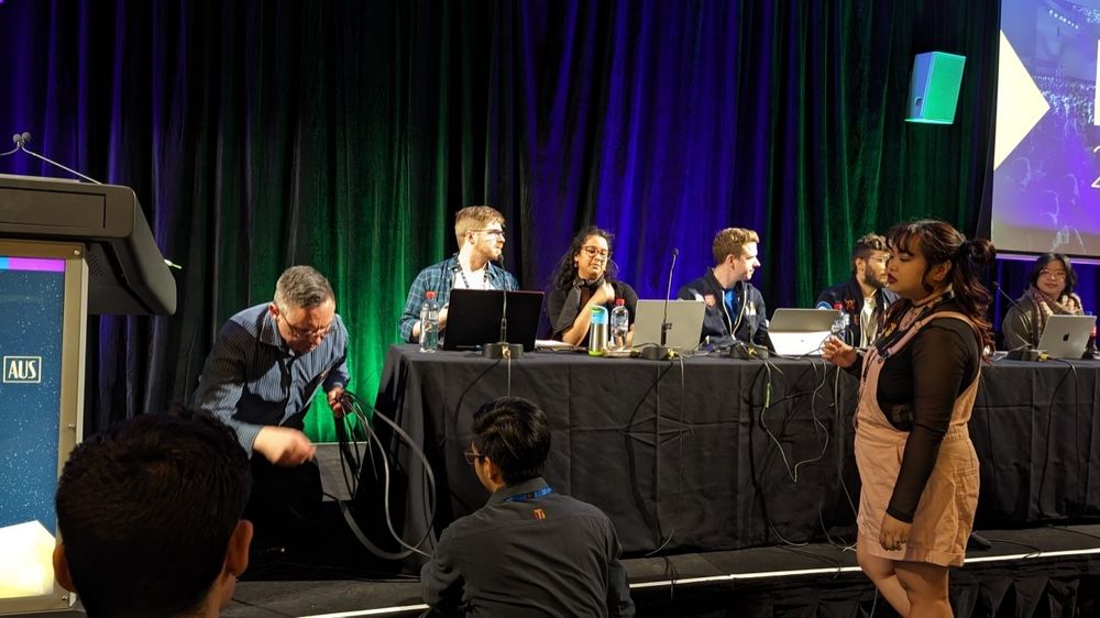 A panel of six people sitting behind tables crowded with laptops and cables. A man kneels next to the table, a knot of cables in his hand.