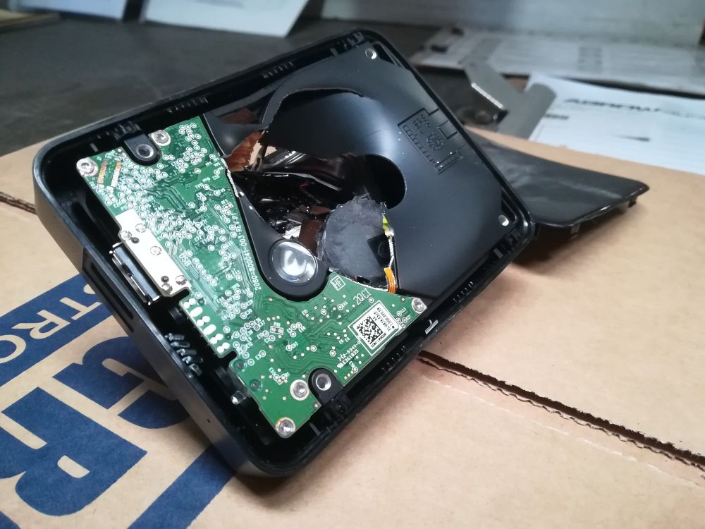 The top of a portable hard drive. The cover has popped off, revealing the inner workings. A large circular divot is visible in the remaining plastic. The green circuit board has been snapped in half.