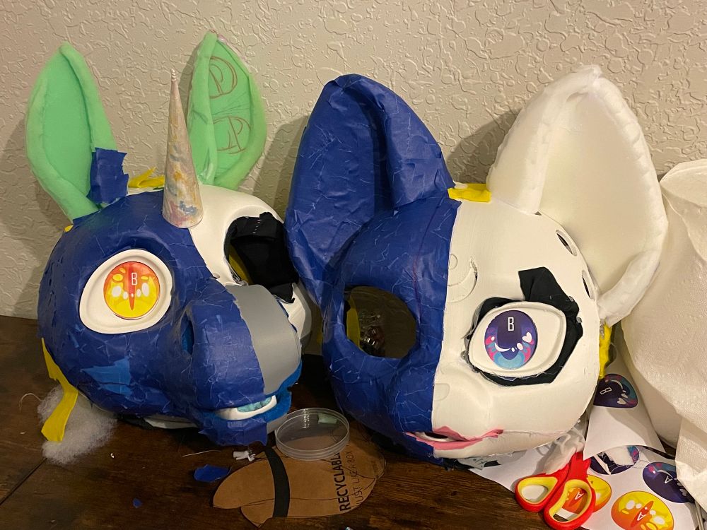 Two work in progress (WIP) fursuit heads. The fursuit head on the left is a unicorn with a gold eye, the one on the right is a cat with a blue eye. Both suits are 3D printed and taped with blue painters tape. 