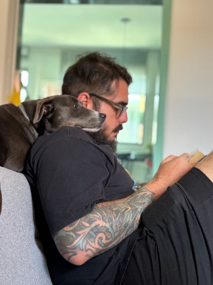pedrotpo.bsky.social sentado no sofá lendo um livro, enquanto sua cachorrinha cinza, deitada na parte superior do sofá, apoia a cabeça no ombro de Pedro, dando a impressão de estar lendo o livro junto ao seu tutor