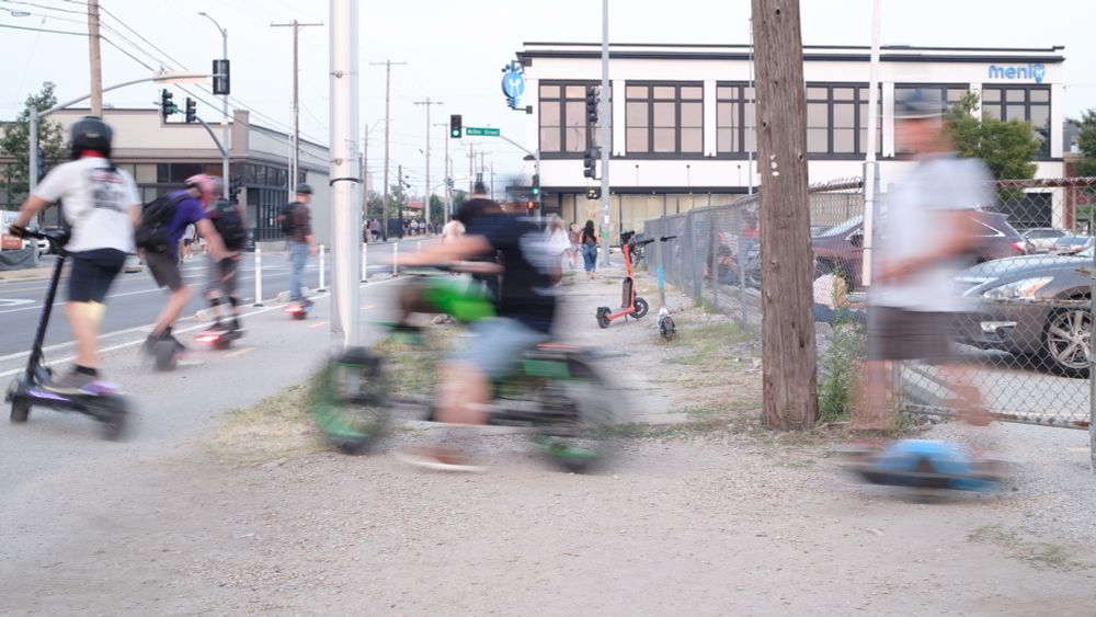 Several people on electric scooters/bikes exit a parking lot in a single file line turning down the street. 