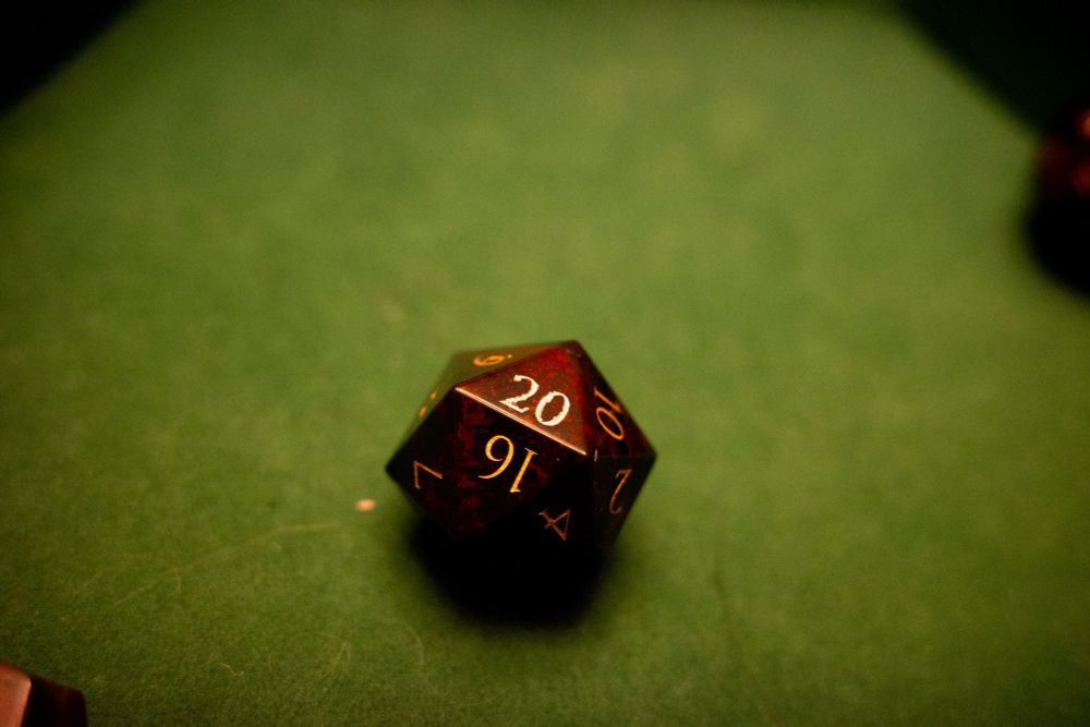 A dark red 20 sided dice with gold leaf numbers resta on a carpet of green felt. 