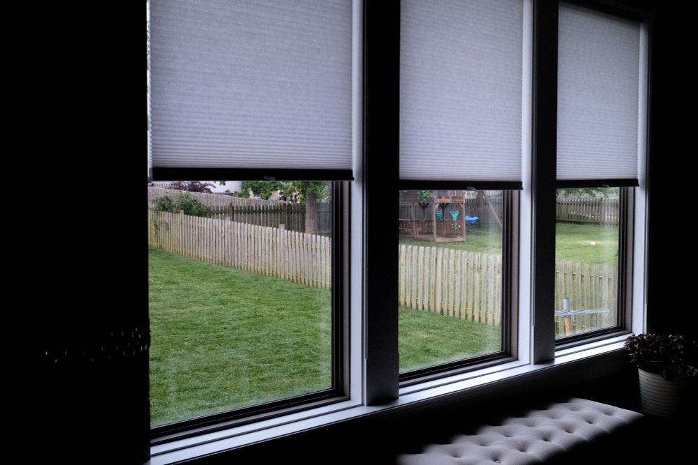 a soccer ball sits in a small spot of sunlight in the far distance of a near by set of windows. The inside of the room is pitched in darkness and only small details are visible from a small bench and a flower pot. The flowers in the flower pot died long ago, and only the stems of their former flowers are left. Looking through the windows you can see a low picket fence splitting two yards. there is a wodden play set in the neighboring yard, along with soccer ball. The grass is green and recently cut in both yards. 