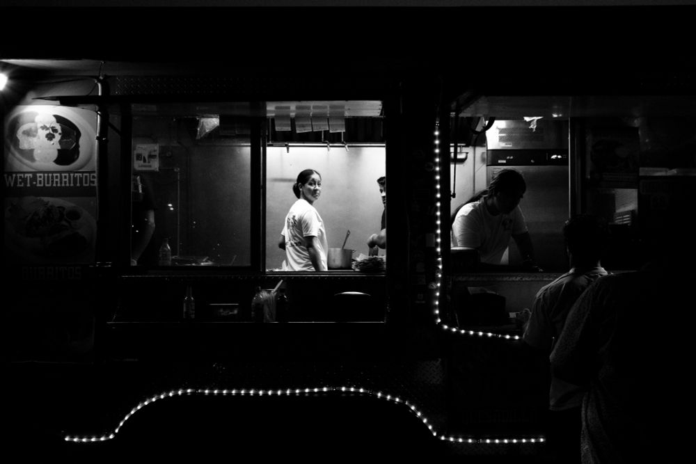 A black and white photo of a food truck with a working looking back out towards the street from one of the order window. 