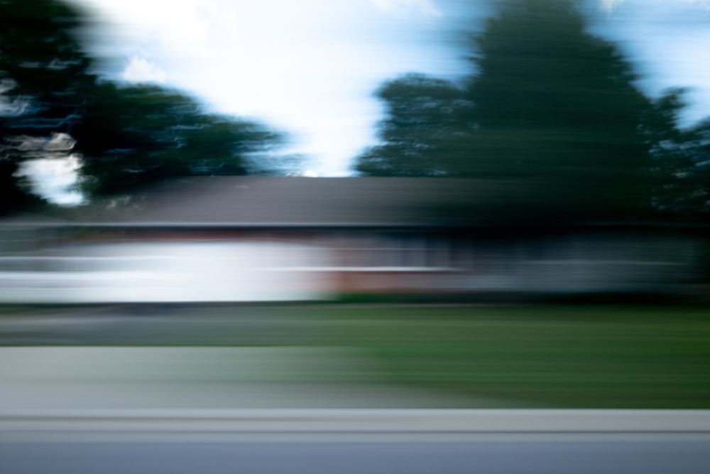 A photograph of a house blurred from left to right. The photo was a long exposure while driving down the street. Trees and a drive way is suggested along with the house. 