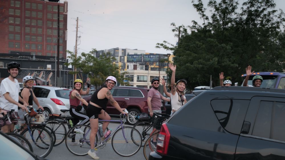 A group of people on bicycles wave to someone off camera in a busy city street. 