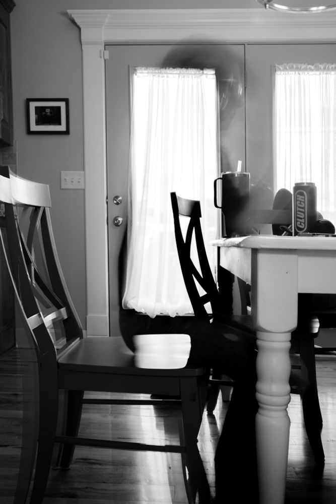 A long exposure photograph of a room with a kitchen table and several chairs with two large window doors in the back ground. There are hints at the movement of a person in the foreground of the image. The chair closets to the viewer shows movement from the person standing up from the table. Only a small part of the table is visible. It is mostly cut off on the right side. 